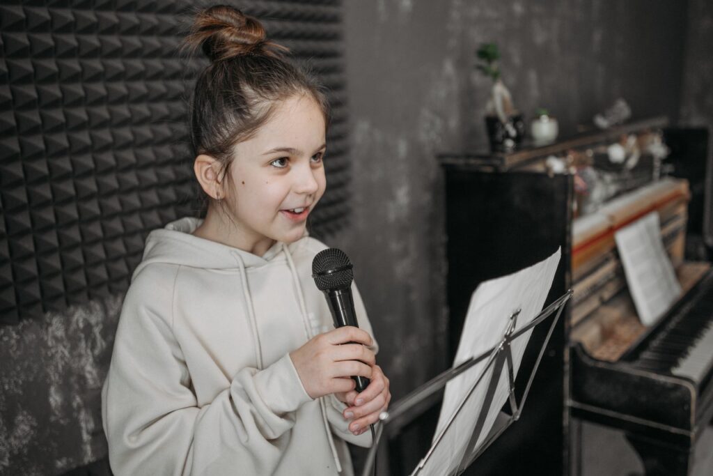 pexels photo 7521359 Caucasian girl singing into a microphone in a soundproof studio with sheet music and piano.